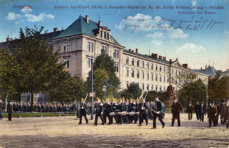 Vor einem langgestrecktem vierstöckigen Gebäude spielt eine Militärkapelle, während weitere Soldaten im Hintergrund zuschauen.