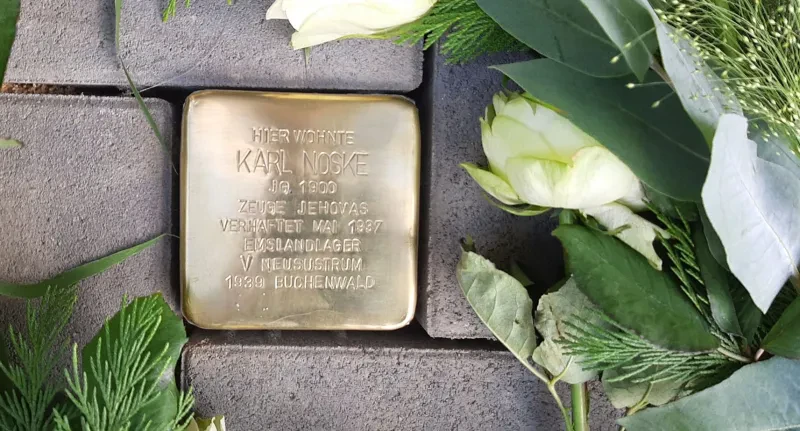 Ein frisch verlegter Stolperstein mit dem Text: "Hier wohnte Karl Noske, Jahrgang 1900, Zeuge Jehovas, verhaftet Mai 1937, Emslandlager V Neusustrum, 1939 Buchenwald". Neben dem Stein liegen weiße Rosen.