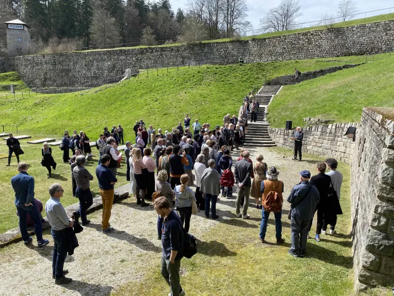 Eine Menschenmenge vor einer Steinmauer, ein Redner steht vor einem Mikrofon, hinter ihm eine noch zu großen Teilen verdeckte Gedenktafel. Im Hintergrund ein Wachtturm.