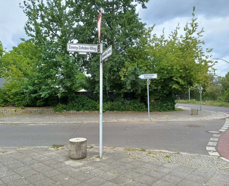 Eine Straßeneinmündung mit dem Straßenschild „Emmy-Zehden-Weg“. Auf der gegenüberliegenden Straßenseite befindet sich ein Hinweisschild mit der Aufschrift „Gedenkstätte Plötzensee“.