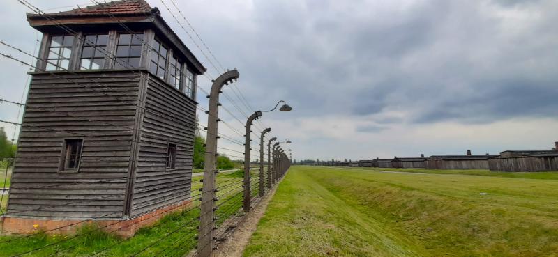 Eine Grünfläche mit Graben trennt einen Elektrozaun von einer Reihe aus 18 Holzbaracken. Hinter dem Zaun steht ein Wachturm.