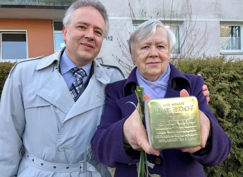 Zwei Personen im Wintermantel stehen vor einem Wohnhaus und halten einen Stolperstein hoch, der die Aufschrift trägt: "Hier wohnte Ludwig Bischoff, Jahrgang 1900, Zeuge Jehovas, verhaftet 12.9.1936, Columbia-Haus, Berlin, 1936 Sachsenhausen, Gefängnis Berlin-Tegel, entlassen 29.3.1938."