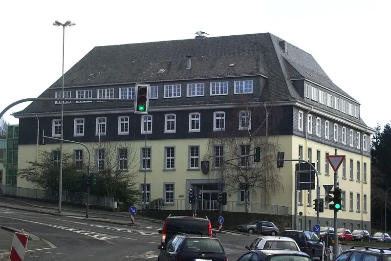 Ein vierstöckiges Gebäude mit Walmdach steht an einer belebten Ampelkreuzung. Die unteren beiden Stockwerke sind hellgelb verputzt, während die oberen beiden Stockwerke dunkel verschiefert sind.