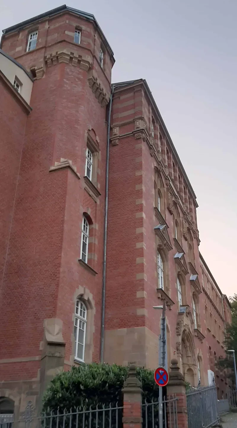 An einer Straßeneinmündung befindet sich ein vierstöckiges, L-förmiges Gebäude aus roten Ziegeln, dessen Knickstelle von einem Turm gebildet wird.