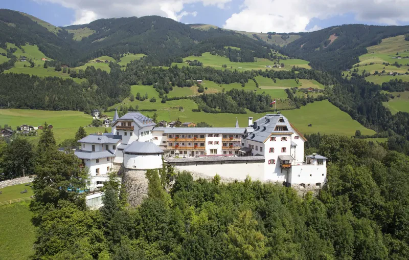 Ein Schloss auf einem Berg im Grünen, dahinter Berge.