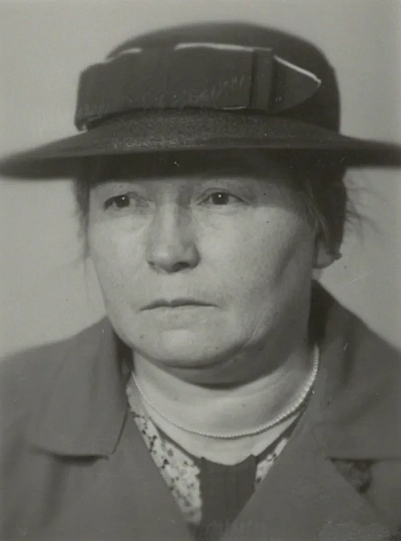 Eine ältere Dame mit nachdenklichem Blick. Sie trägt einen schicken Hut und eine Perlenkette.