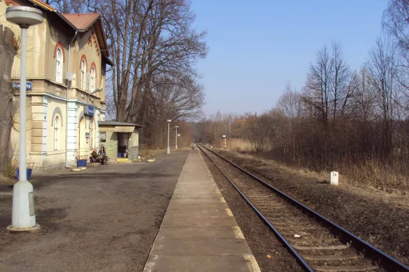 Schienen, links ein Bahnhofsgebäude, zwei Personen sitzen davor.