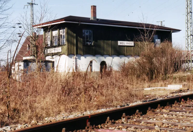 Ein Bahnhofsgebäude hinter Schienen und Gestrüpp.