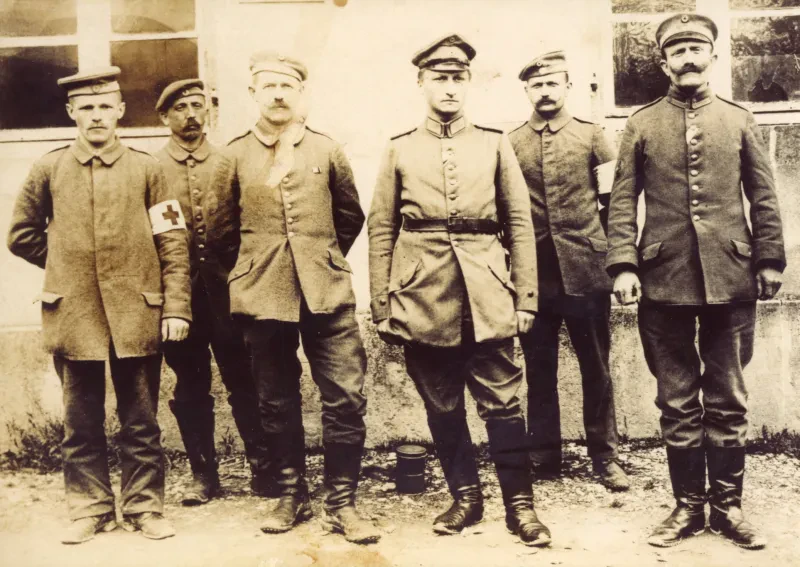 Fünf Soldaten in Uniformen in einer Reihe vor einem Haus. Der links außen stehende trägt eine Rot-Kreuz-Binde.