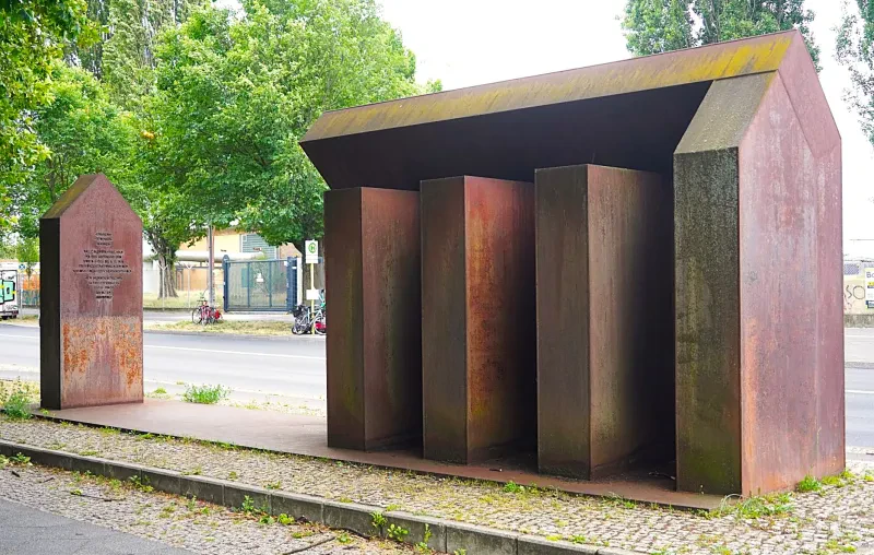 Vier aufrecht stehende Stahlquader stellen ein offenes Haus dar, dass auf der Rückseite zur Straße hin durch eine weitere Stahlplatte geschlossen ist. In kurzer Distanz befindet sich eine weitere Stahlwand mit einer Inschrift.