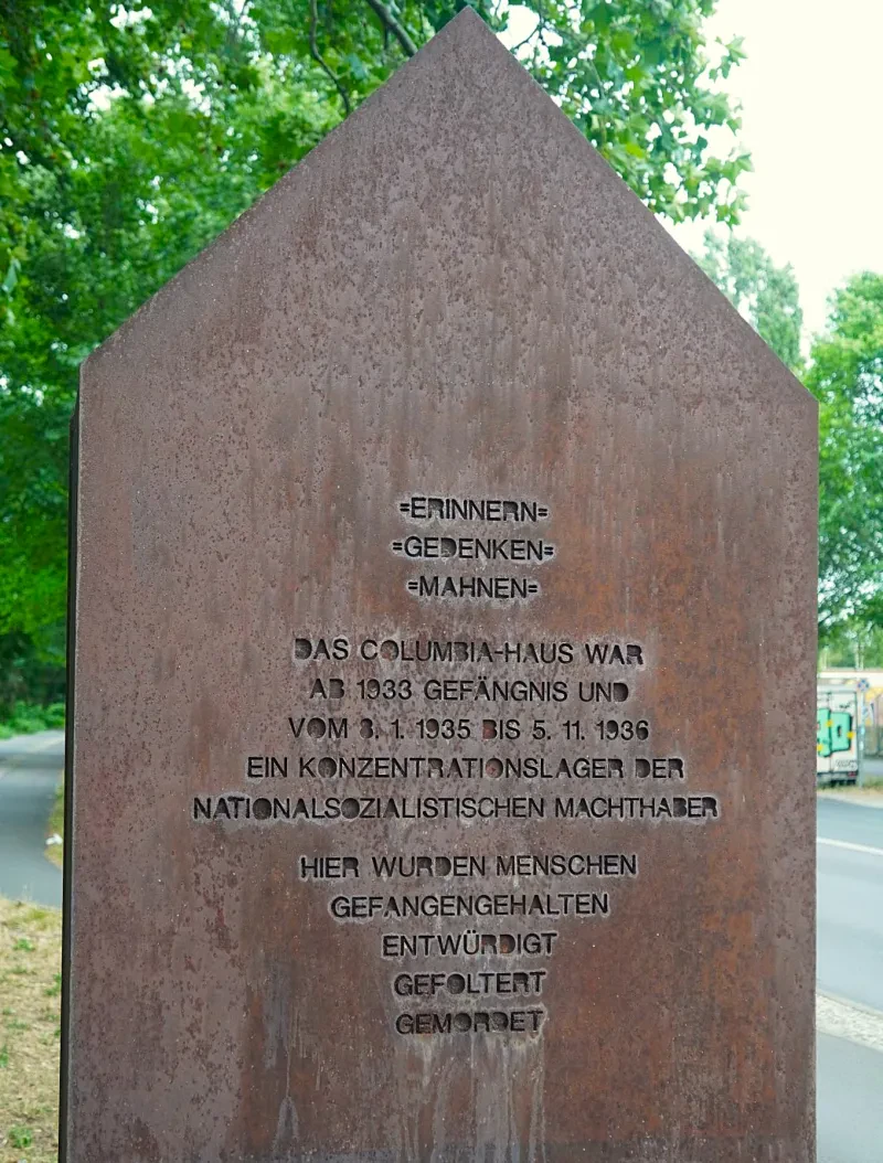 Stilisierte Giebelwand aus Stahl mit der Inschrift: „Erinnern / Gedenken / Mahnen / Das Columbia-Haus war ab 1933 Gefängnis und vom 8.1.1935 bis 5.1.1936 ein Konzentrationslager der nationalsozialistischen Machthaber. Hier wurden Menschen gefangengehalten, entwürdigt, gefoltert, gemordet.“