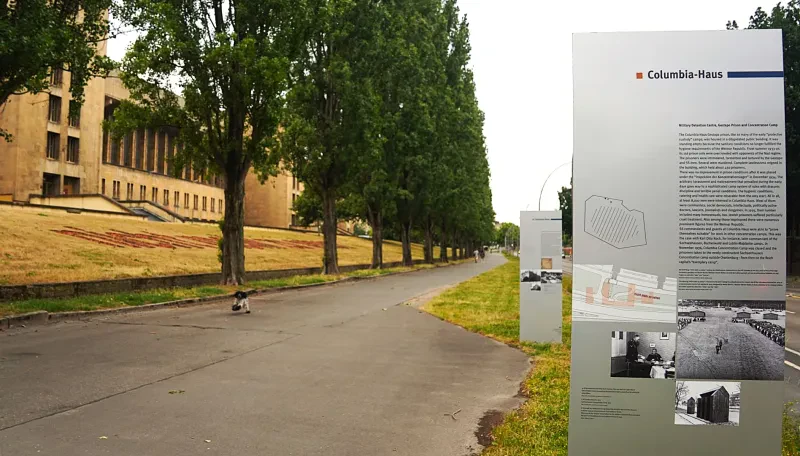 Zwei Schautafeln mit zweisprachigem Text in Deutsch und Englisch, sowie mit Fotos und Kartenausschnitten stehen an einem breiten Fußweg zur Straßenseite. Auf der anderen Seite des Fußwegs sieht mit hinter einer Baumreihe einen Teil der Abfertigungshalle des ehemaligen Flughafens.