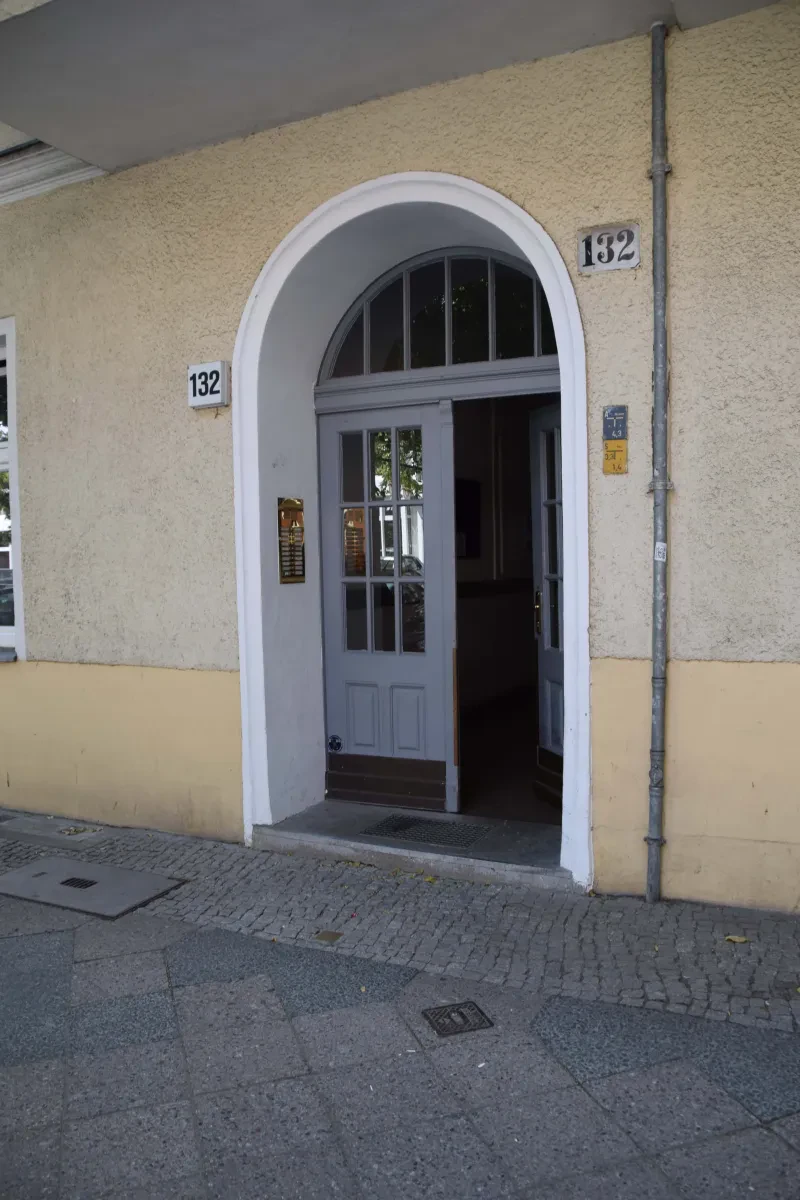 Ein Hauseingang, links ist die messingglänzende Klingelanlage zu sehen. Die Tür steht offen. Neben dem Eingang befindet sich oben rechts und links die Hausnummer 132. Vor dem Eingang ist im Boden ein Stolperstein zu sehen.