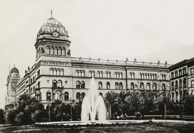 Ein von einer Grünanlage umgebener Springbrunnen steht vor der Fassade eines vierstöckigen Prungebäudes mit gewölbten Fenstern und Ecktürmen mit Uhr.