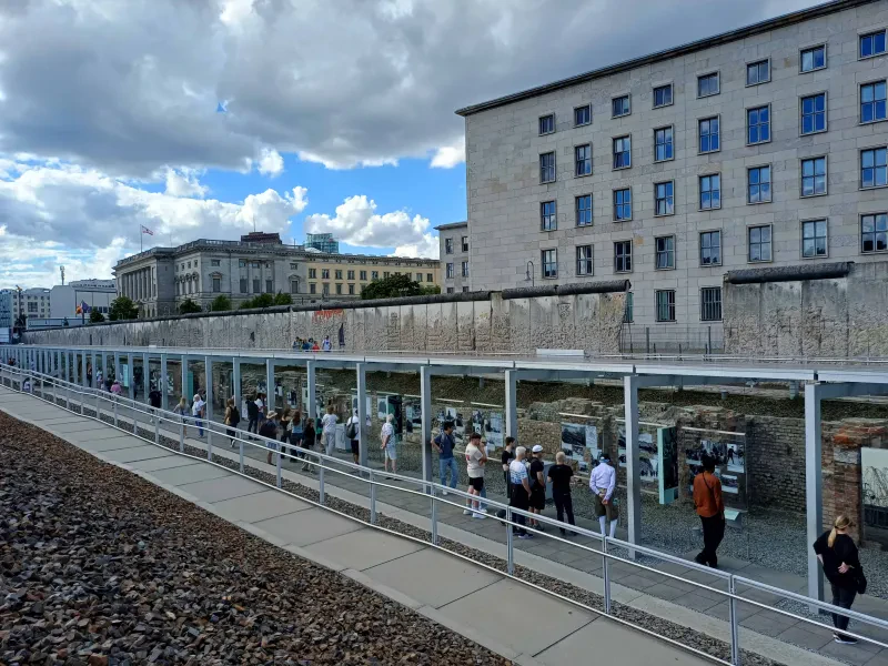 Menschen sehen sich eine überdachte Open-Air-Ausstellung an, im Hintergrund sind Reste der Berliner Mauer.