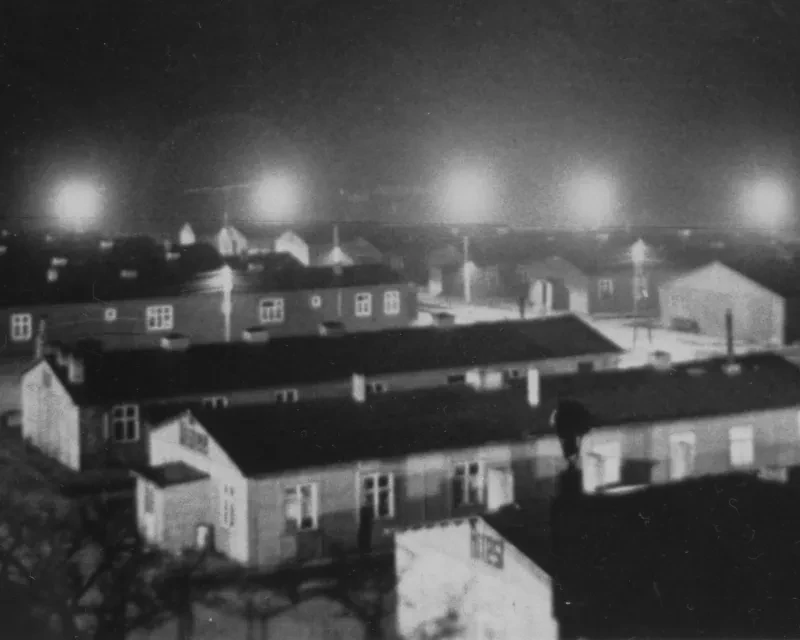 Scheinwerfer erhellen das Lager bei Nacht. Die Holzbaracken haben auf der Längsseite gewöhnlich sieben Fenster. (Bildausschnitt)