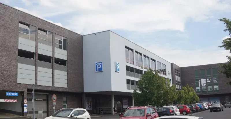 Ein dreistöckiges Einkaufszentrum hell verputzt mit rotbraunen Klinkersteinen. Davor befindet sich ein großer Parkplatz.