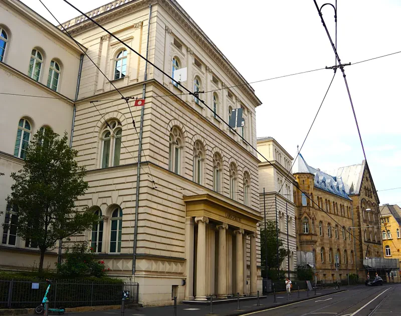 Ein helles, dreistöckiges Prunkgebäude aus großen Steinquadern mit einem Säulenportikus aus dem 19. Jahrhundert steht neben einem dunklen, dreistöckigen Gebäude aus dem beginnenden 20. Jahrhundert.