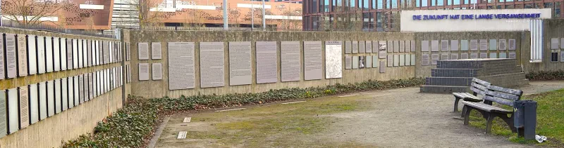 Auf einer langen Betonmauer hängen in zwei Reihen übereinander eine große Zahl von Gedenktafeln.