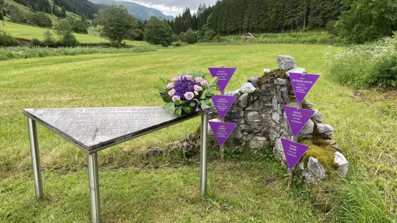 Gedenktafel für die Opfergruppe der Zeugen Jehovas in der Gedenkstätte KZ-Nebenlager Bretstein, 2023. Als Tisch aufgestelltes Dreieck und sechs lila Winkel an dem Rest einer Steinmauer, im Hintergrund eine Wiese, Wald und Berge. (Verein Lila Winkel, Fotografin: Heide Gsell).