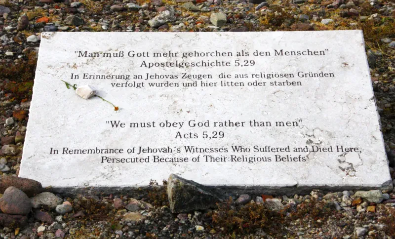 Auf einer hellen Gedenktafel stehen in dunkler Schrift in deutscher und englischer Sprache die Worte: "Man muß Gott mehr gehorchen als den Menschen" - Apostelgeschichte 5, 29 - In Erinnerung an Jehovas Zeugen die aus religiösen Gründen verfolgt wurden und hier litten oder starben".