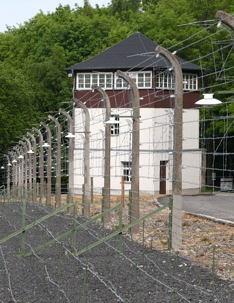 Hinter einem elektrisch geladenen und einem Stacheldrahtzaun steht ein dreistöckiger Wachturm mit breiten Fenstern und mit spitzem Dach.
