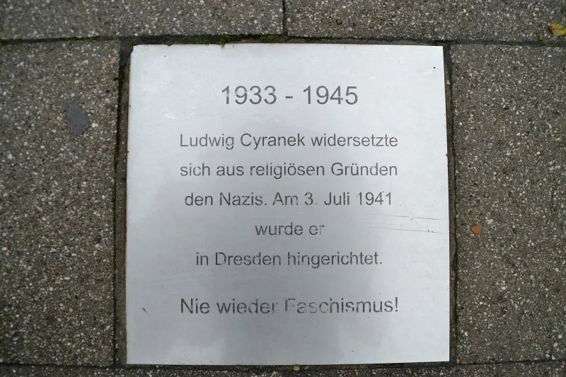 Auf einer silbernen Gedenkplatte steht der Text "1933 – 1945 Ludwig Cyranek widersetzte sich aus religiösen Gründen den Nazis. Am 03. Juli 1941 wurde er in Dresden hingerichtet. Nie wieder Faschismus!"