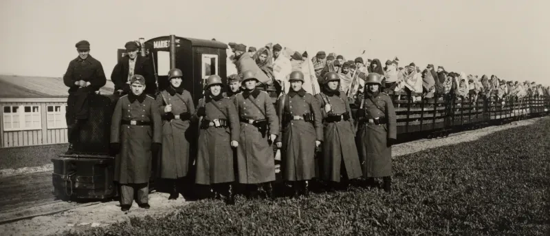 Eine kleine Lokomotive zieht offene Wagons, die mit vielen Männern überladen sind. Davor stehen acht Soldaten in NS-Uniform und zwei Lokführer.