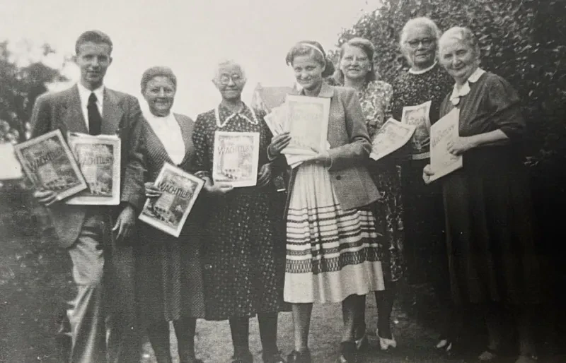 Ein junges Paar und fünf ältere Frauen posieren in einem Garten mit der der Zeitschrift "Der Wachtturm" in der Hand.
