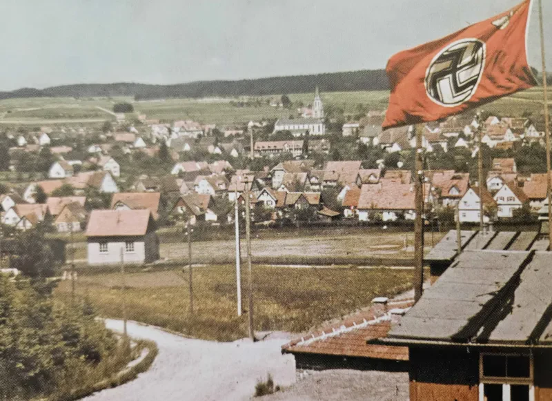 Blick auf eine Wiese, dahinter ein Dorf, im Vordergrund weht eine Hakenkreuzfahne.