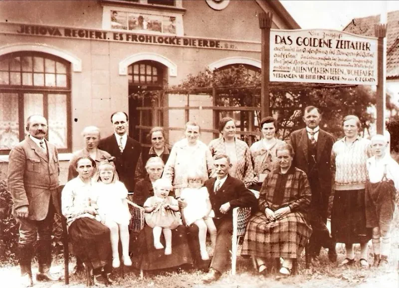 Eine Gruppe von Männern, Frauen und Kindern sitzend und stehend vor einem Haus, an dessen Giebel "Jehova regiert. es frohlockt die Erde." steht. Ein Schild macht Werbung für die Zeitschrift "Das Goldene Zeitalter".