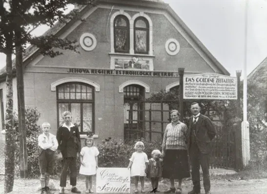 Das Ehepaar Fokken und fünf ihrer Kinder stehend vor dem Haus der Familie, an dessen Giebel "Jehova regiert. es frohlockt die Erde." steht. Ein Schild macht Werbung für die Zeitschrift "Das Goldene Zeitalter". Zwei Mädchen halten ein Schild mit der Aufschrift "Eine glückliche Familie".