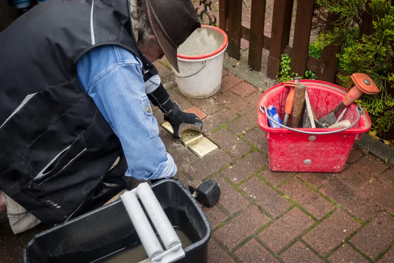 Ein Mann in Arbeitskleidung und mit Hut verlegt drei Stolpersteine. Er setzt den mittleren Stein in den Gehweg.