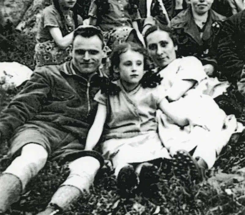 Eine Familie sitzt im Gras, Vater links, Tochter mitte, Mutter rechts. Hinter ihr sitzen weitere Personen. Es sieht aus, als würden sie sich auf einem Ausflug befinden.