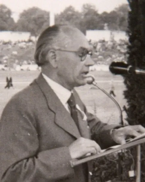Ein älterer Mann vor einem Mikrofon, er stützt sich auf ein Pult. Im Hintergrund ist ein Stadion zu erkennen.