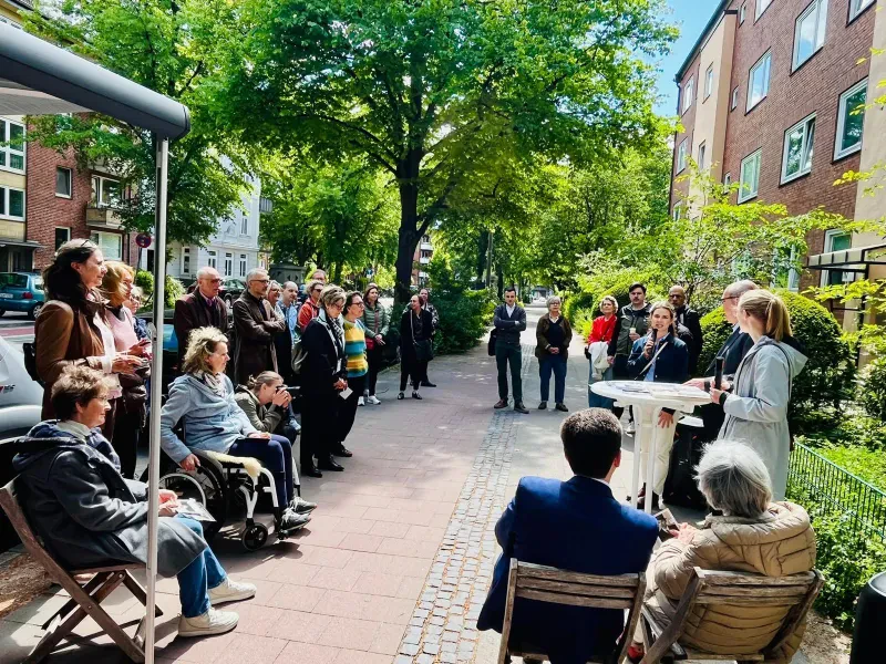 Die Veranstaltung DENK MAL AM ORT. Drei Menschen stehen an einem Tisch vor einem Hauseingang. Ein größere Gruppe von Menschen steht im Halbkreis um sie herum.
