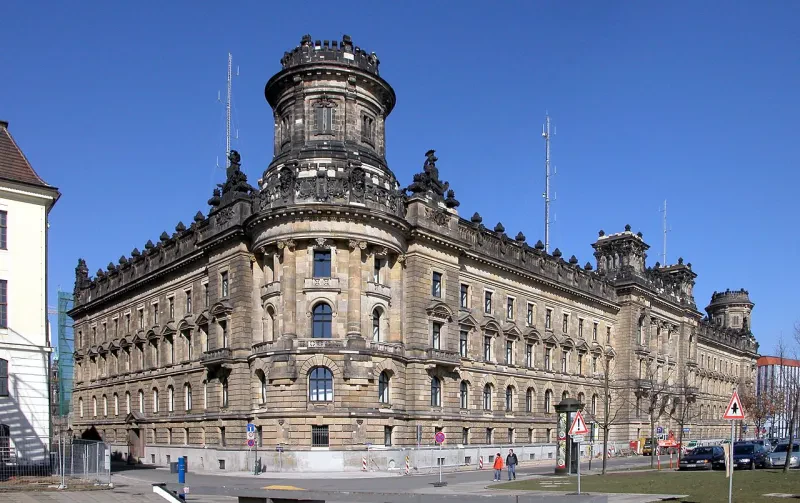 Ein langezogenes sandsteinfarbenes Gebäude mit Dachballustrade steht repräsentativ an einem öffentlichen Platz.
