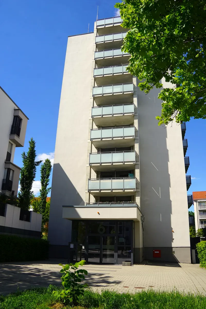 Ein elfstöckiges, hell verputztes Wohnhaus mit dunklen Aluminiumtüren und hellblauen Balkons zur Straßenseite.