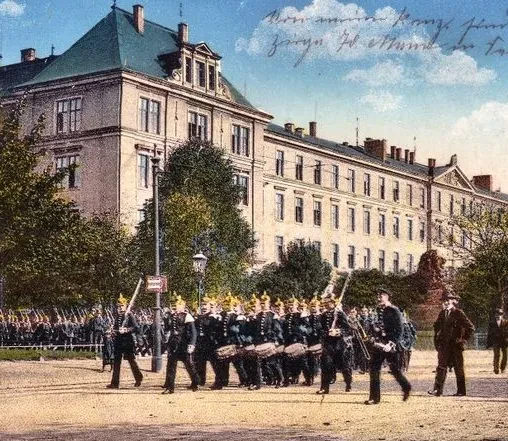 Vor einem langgestrecktem vierstöckigen Gebäude spielt eine Militärkapelle, während weitere Soldaten im Hintergrund zuschauen.