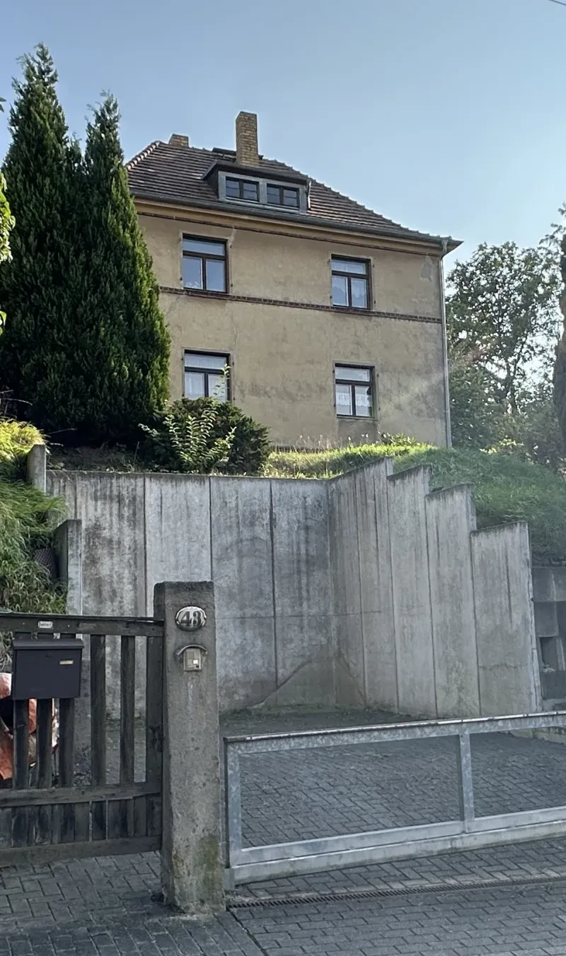 Auf einer Anhöhe steht ein dreistöckiges Wohnhaus, dessen oberstes Stockwerk im Walmdach ausgebaut ist.