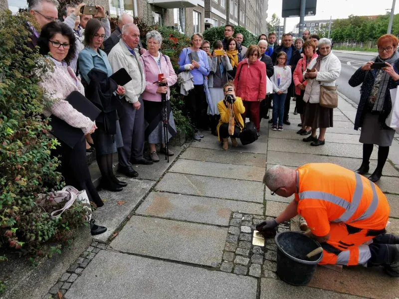 Ein Mitarbeiter der Stadt Dresden reinigt zwei frisch verlegte Stolpersteine. Er wird dabei aufmerksam von 26 Passanten beobachtet, von denen einige Fotos machen.
