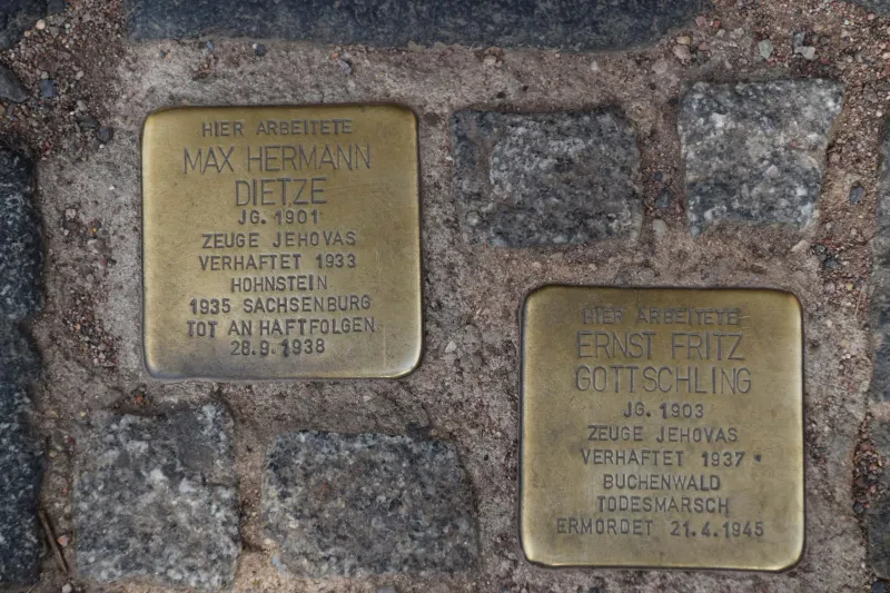 Zwei Stolpersteine mit dem Text "HIER ARBEITETE MAX HERMANN DIETZE, Jahrgang 1901, ZEUGE JEHOVAS, VERHAFTET 1933, HOHNSTEIN, 1935 SACHSENBURG, TOT AN HAFTFOLGEN 28.9.1938" und "HIER ARBEITETE ERNST FRITZ GOTTSCHLING, Jahrgang 1903, ZEUGE JEHOVAS, VERHAFTET 1937, BUCHENWALD, TODESMARSCH, ERMORDET 21.4.1945".