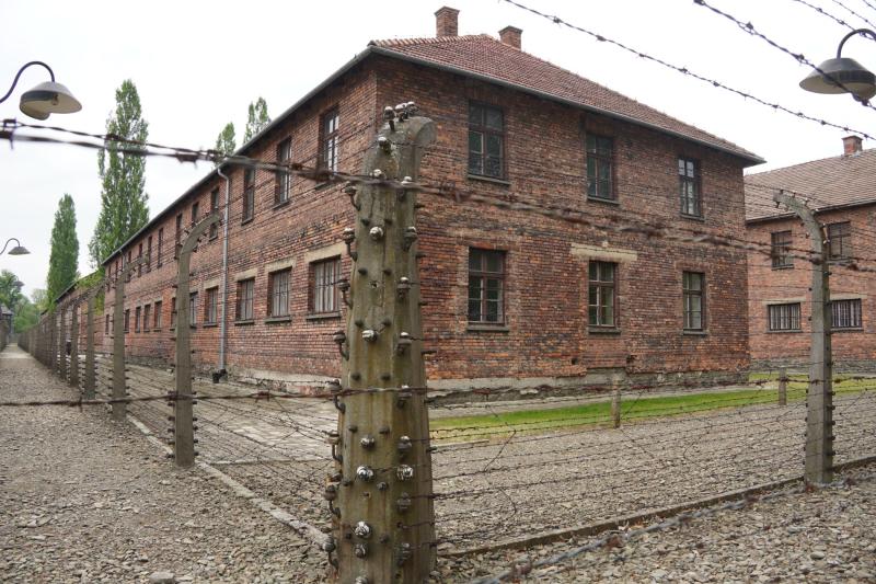 Ein zweistöckiges Gebäude aus roten Backsteinen mit Walmdach steht in der Ecke eines mit Stacheldraht eingezäunten Areals.