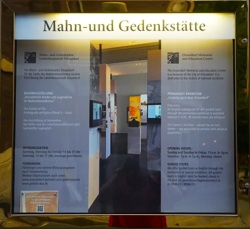 Schaukasten mit der Mitteilung: "Die Mahn- und Gedenkstätte Düsseldorf für die Opfer des Nationalsozialismus ist eine Einrichtung der Landeshauptstadt Düsseldorf." Darunter befinden sich Informationen zur Dauerausstellung, Öffnungszeiten und Führungen in deutscher und englischer Sprache.