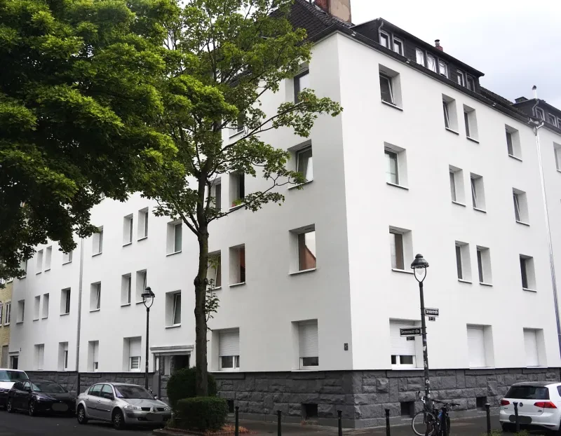 Ein großes, weiß verputztes, fünfstöckiges Eckhaus mit Walmdach und einem Sockel aus Natursteinen.