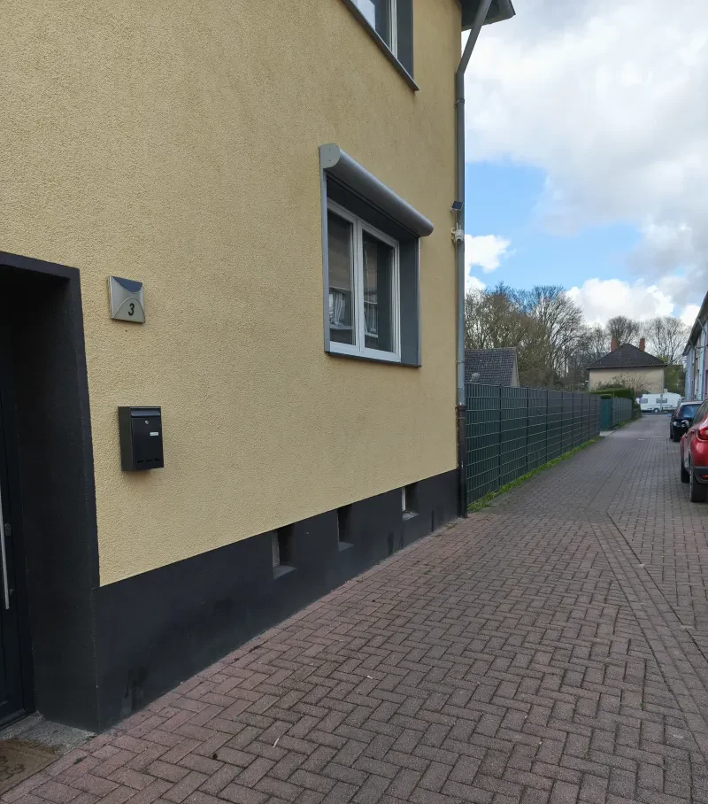 Blick in eine Straße, links steht ein haus, dahinter ein eingezäuntes leeres Grundstück, recht eine reihe parkender Autos.