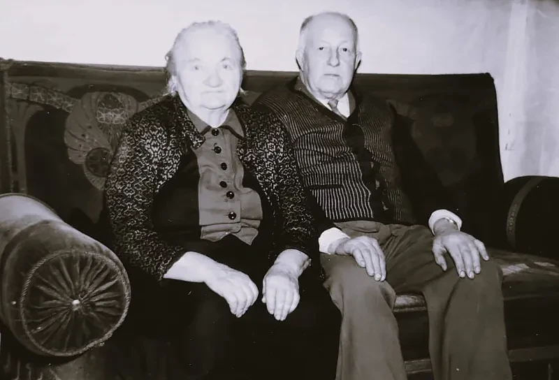 Ein älteres Ehepaar sitzt auf einem Sofa. Sie blicken in die Kamera.