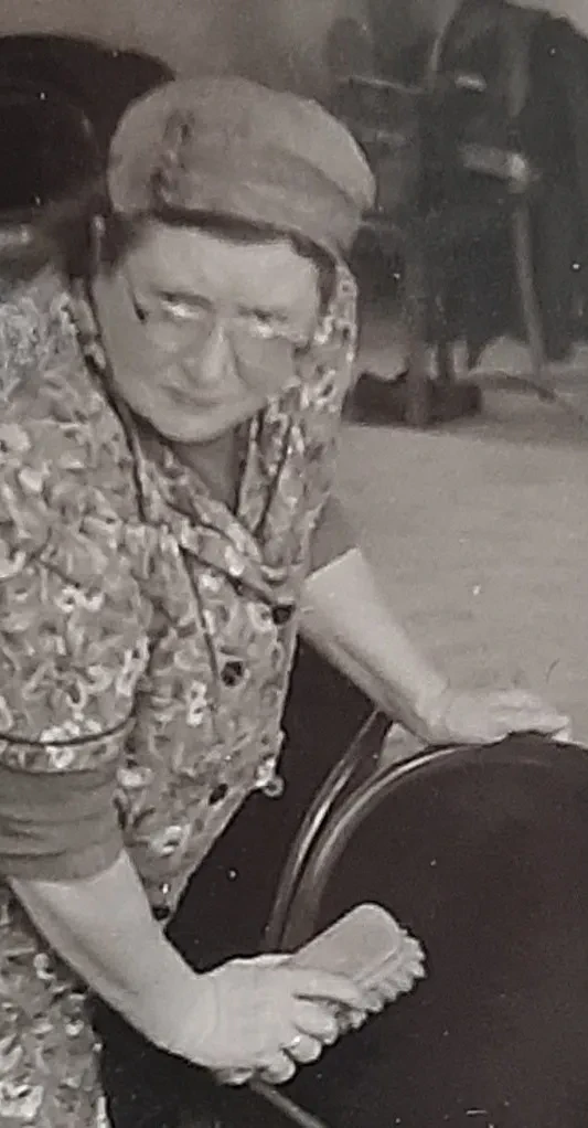 Eine ältere Frau mit Brille und Kittel reinigt Stühle mit einer Bürste.