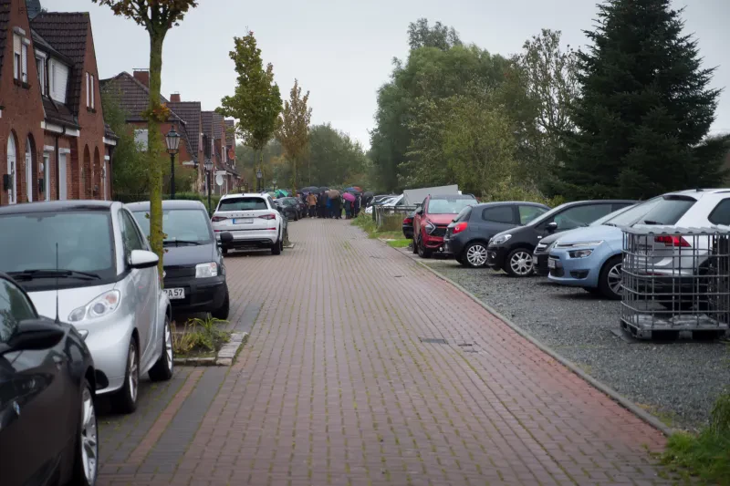Blick in eine Straße, rechts und links stehen Autos, links ist eine Häuserreihe. Am Ende der Straße ist eine Menschenmenge zu sehen, viele tragen Regenschirme.
