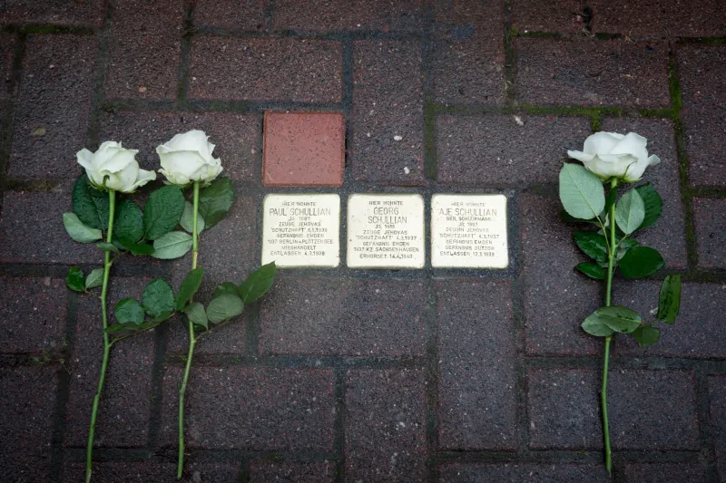 Drei Stolpersteine im Gehweg. Daneben drei Rosen. Die Stolpersteine tragen die Inschriften: "Hier wohnte Paul Schullian / Jg. 1887 / Zeuge Jehovas / 'Schutzhaft' 8.5.1939 / Gefängnis Emden / 1937 Berlin-Plötzensee / Misshandelt / Entlassen 4.3.1939", "Hier wohnte Georg Schullian / Jg. 1918 / Zeuge Jehovas / 'Schutzhaft' 4.3.1937 / Gefängnis Emden / 1937 KZ Sachsenhausen / Ermordet 14.4.1940", "Hier wohnte Aje Schullian geb. Schüürmann / Jg. 1897 / Zeugin Jehovas / 'Schutzhaft' 4.3.1937 / Gefängnis Emden / Gefängnis Bützow / Entlassen 12.5.1938"